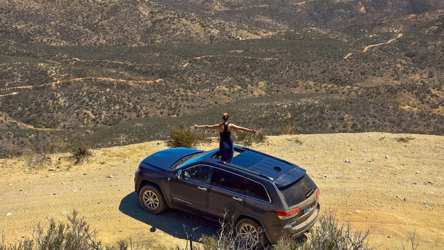A woman stands out of a 2018 Jeep Grand Cherokee atop a mountain's peak.