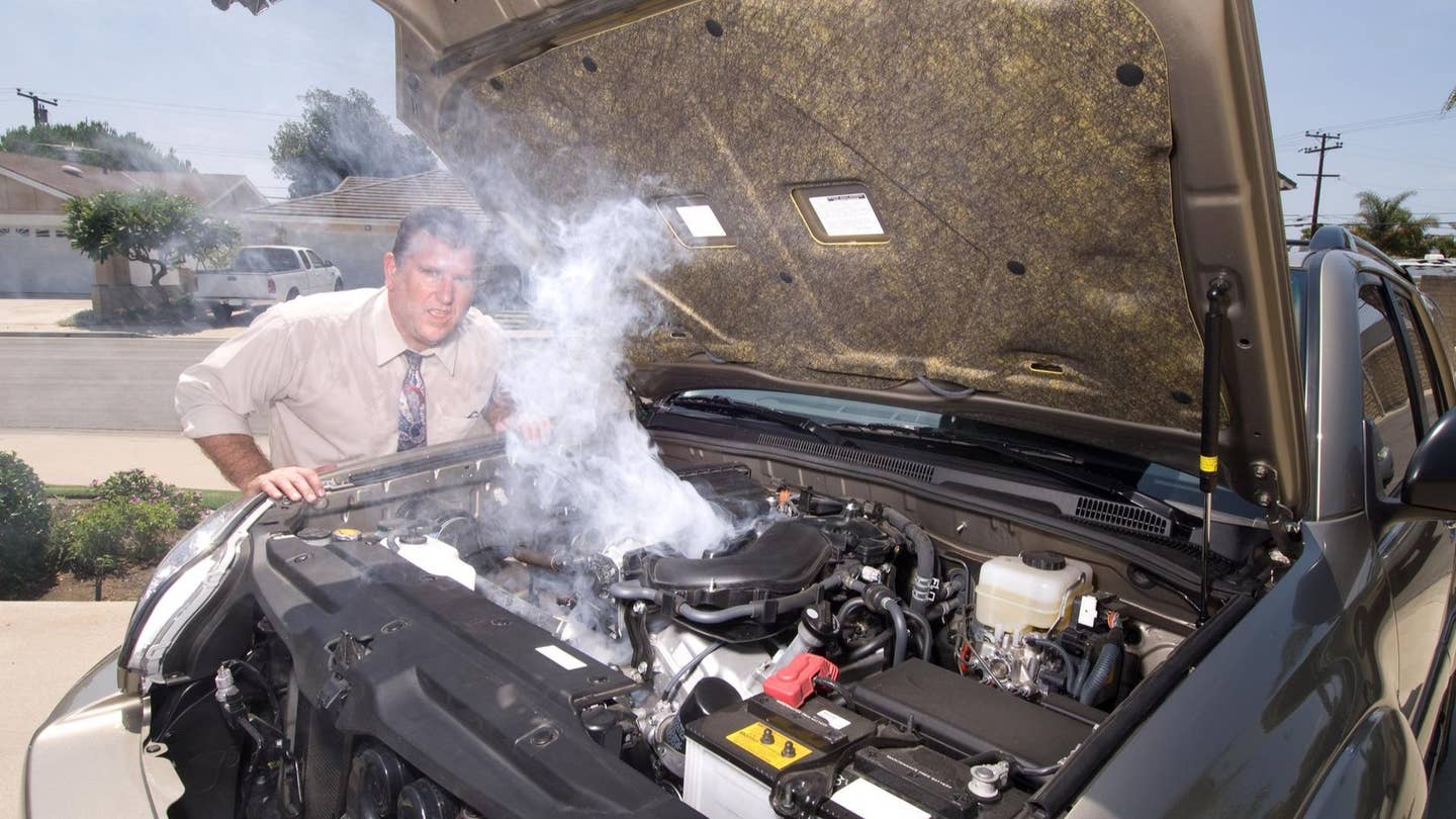 A car's engine smokes as a man looks to camera. 