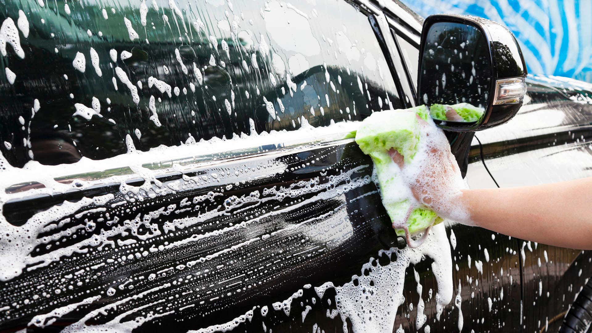 A hand using a sponge to wipe a soapy car door.