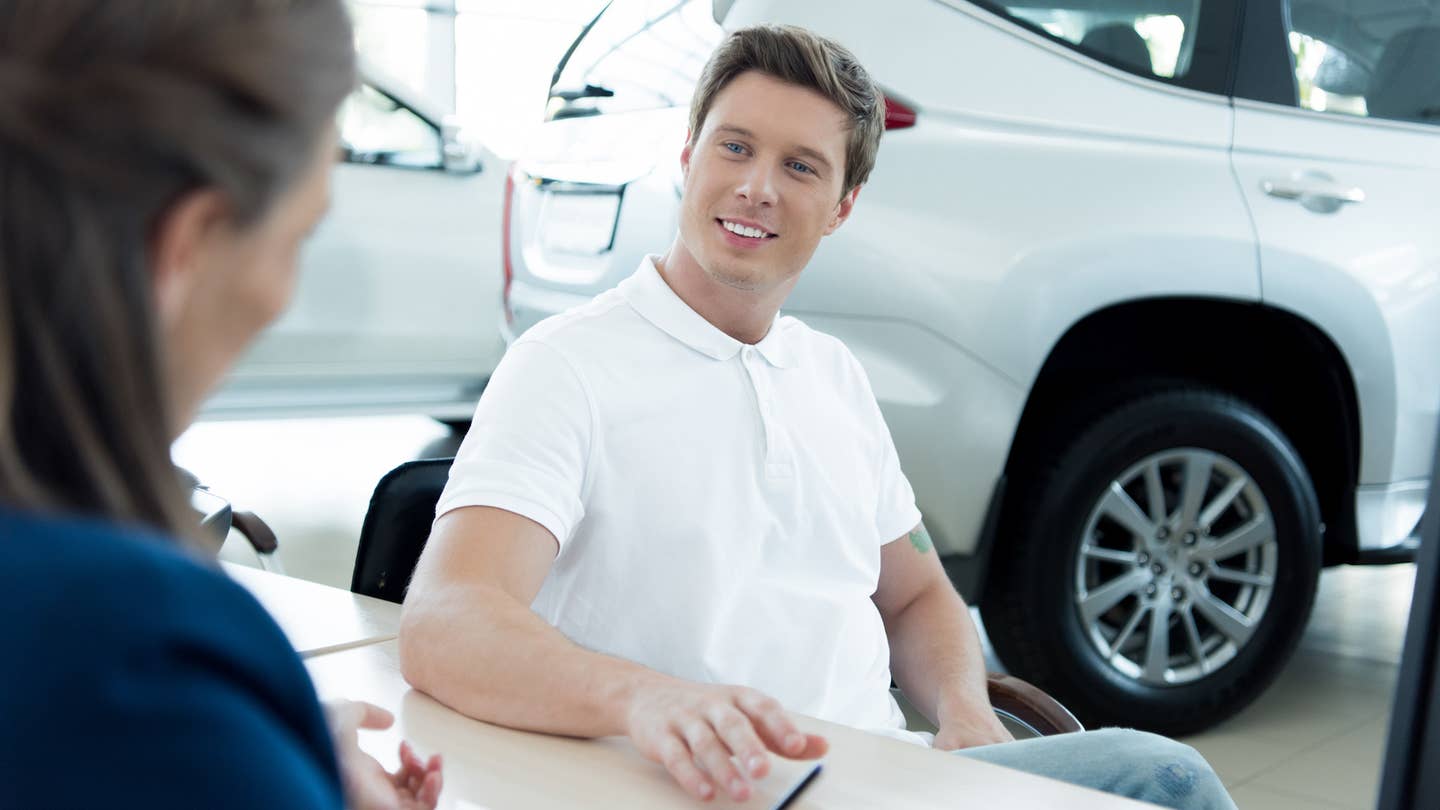 A prospective male buyer in a white polo sits at a car dealership desk.