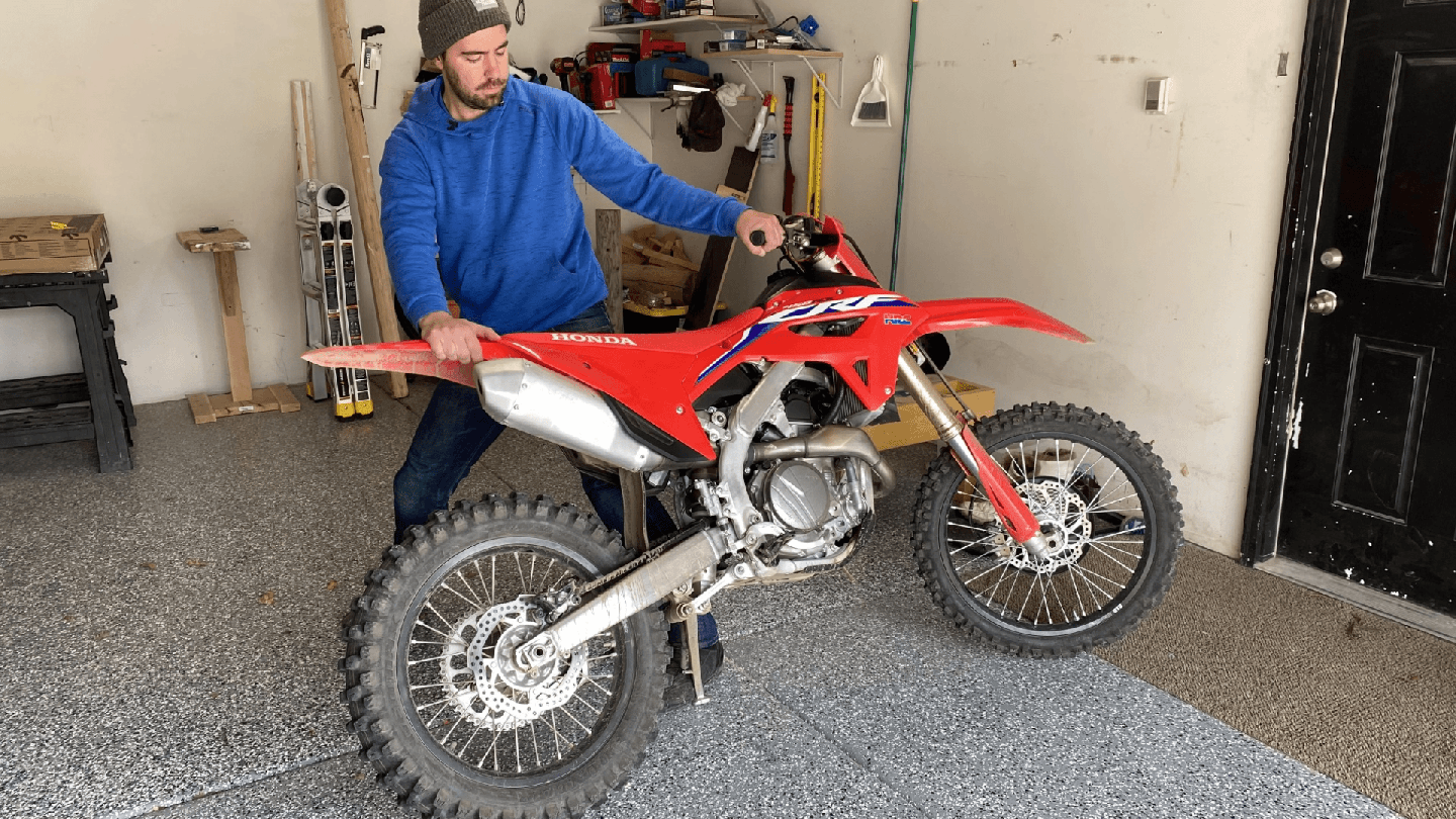 A man rotating a red Honda dirtbike on its kickstand inside a garage.