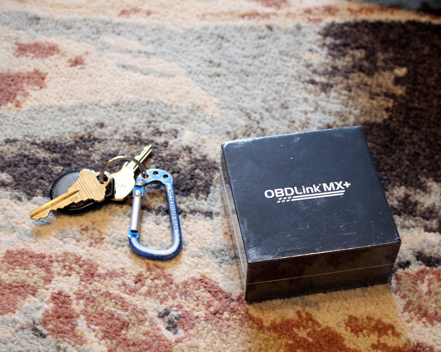 An OBDLink MX+ black box next to keys on top of a multi-colored carpet.