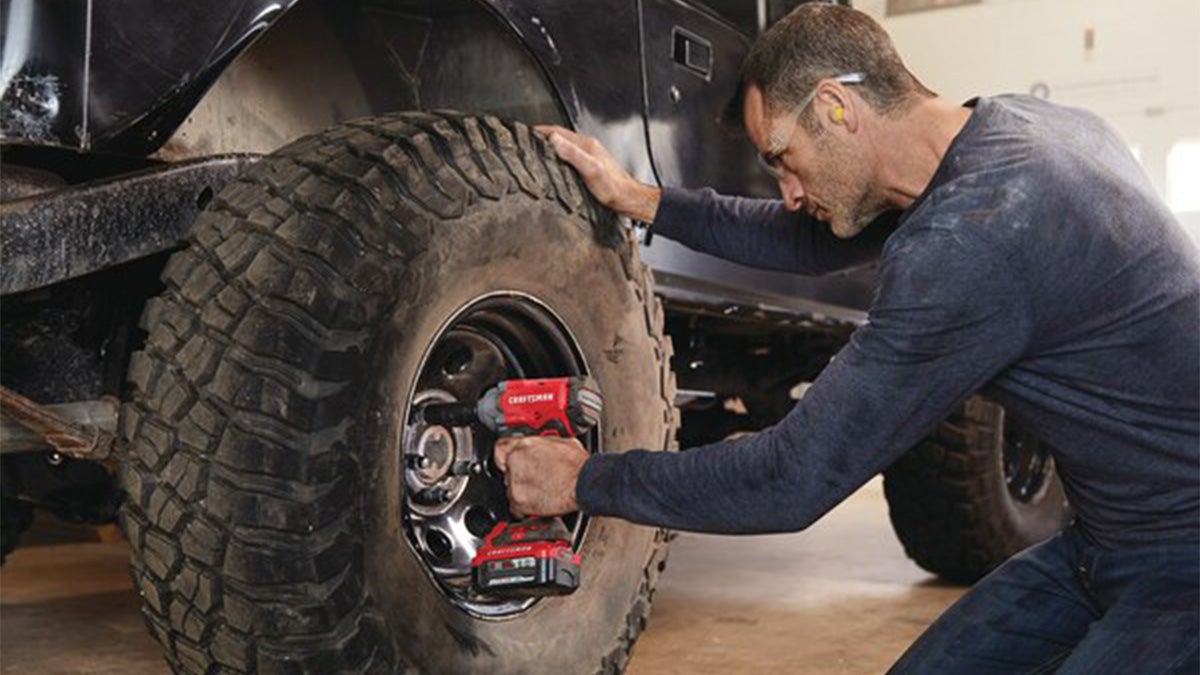 A man in a long sleeve shirt and safety glasses uses an impact wrench to turn lug nuts.