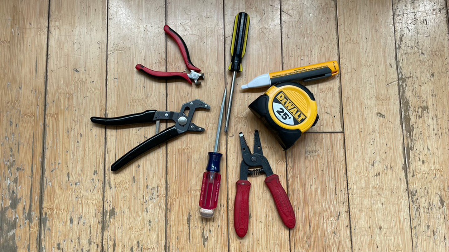 Two pliers, two screwdrivers, a tape measure, an electrical detector, and a crimper arranged in a circle on a wooden floor.