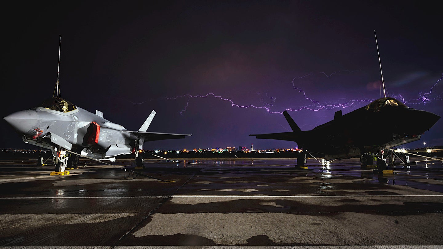 Two US Air Force F-35A Joint Strike Fighters sit underneath lightning rods at Nellis Air Force Base in Nevada while lightning strikes in the background.