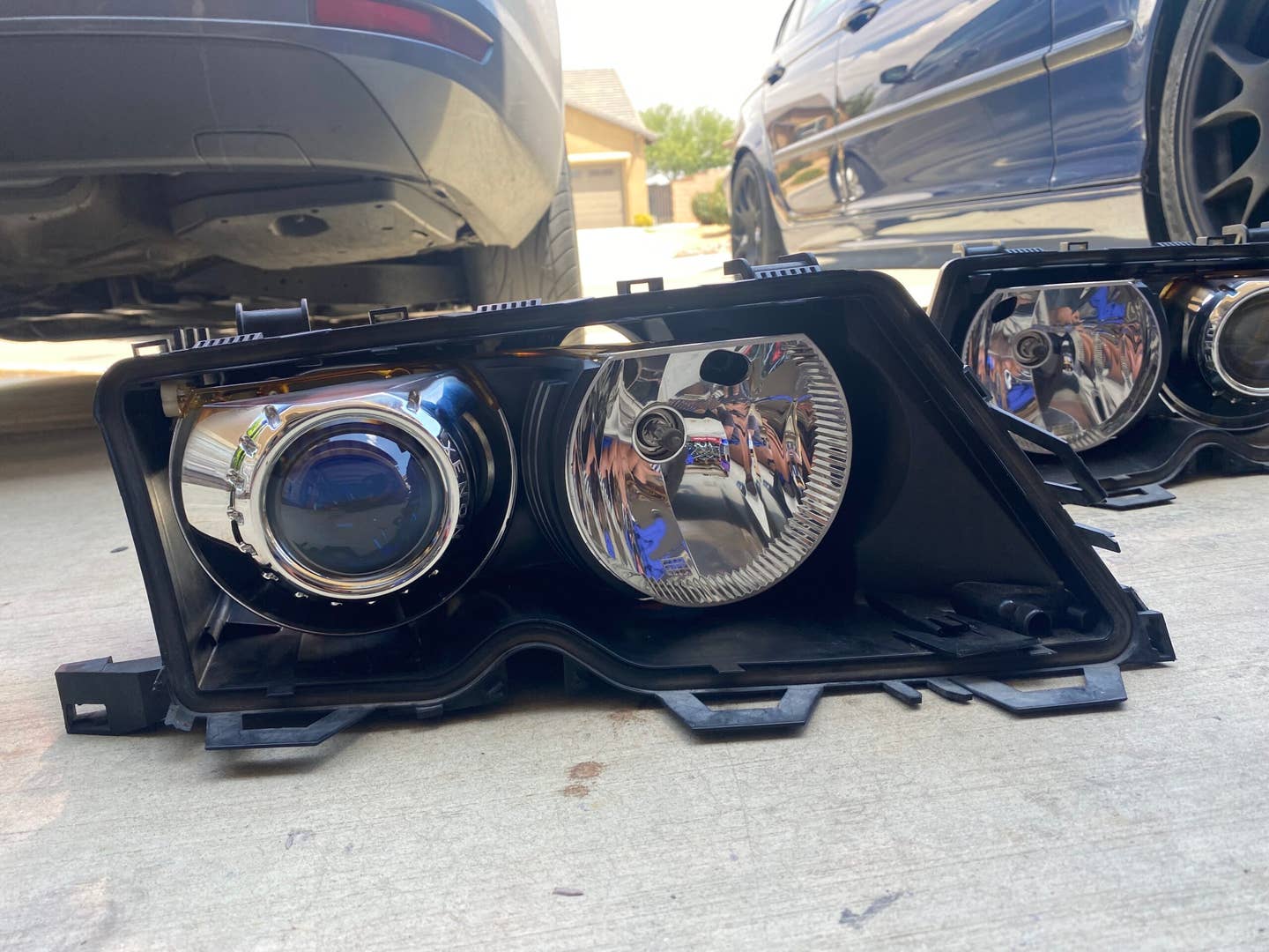A 2004 BMW 330i headlight housing sans lens with new projector retrofit. A blue BMW 330i is off to the right. A grey VW Jetta is to the left.