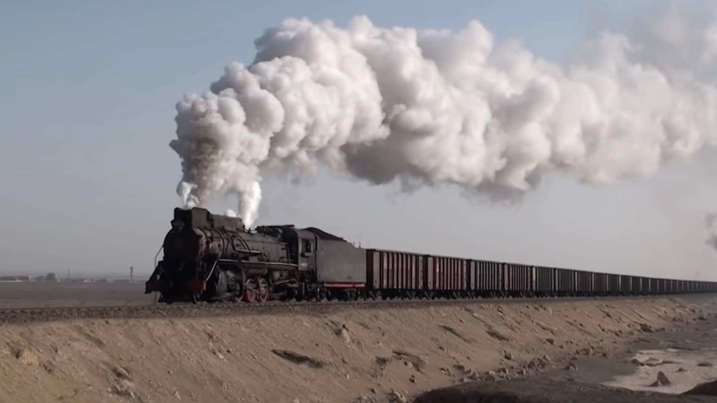 Chinese steam locomotives emitting white clouds of steam and smoke haul coal hoppers against a dry desert backdrop