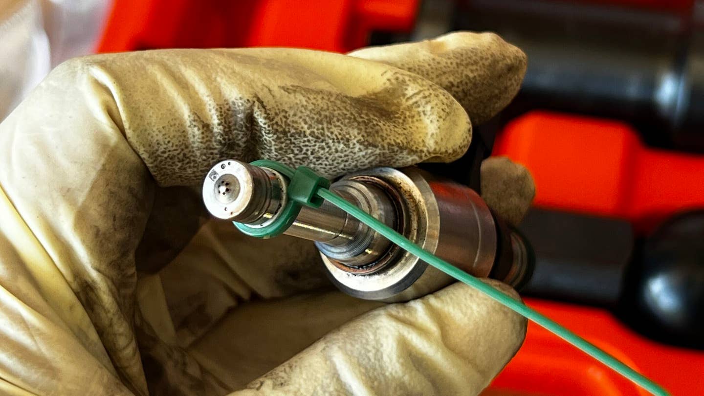 A gloved hand holding a Volkswagen TSI direct injector with the tip facing the camera off to the left. A ziptie is fastened around the white inner seal.