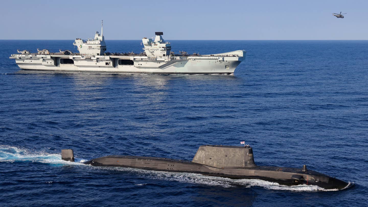 A U.K. Royal Navy&nbsp;<em>Astute&nbsp;</em>class submarine on the surface with the aircraft carrier HMS&nbsp;<em>Queen Elizabeth</em>&nbsp;in the background during the current Carrier Strike Group 21 (CSG21) deployment.&nbsp;<em>Crown Copyright</em>