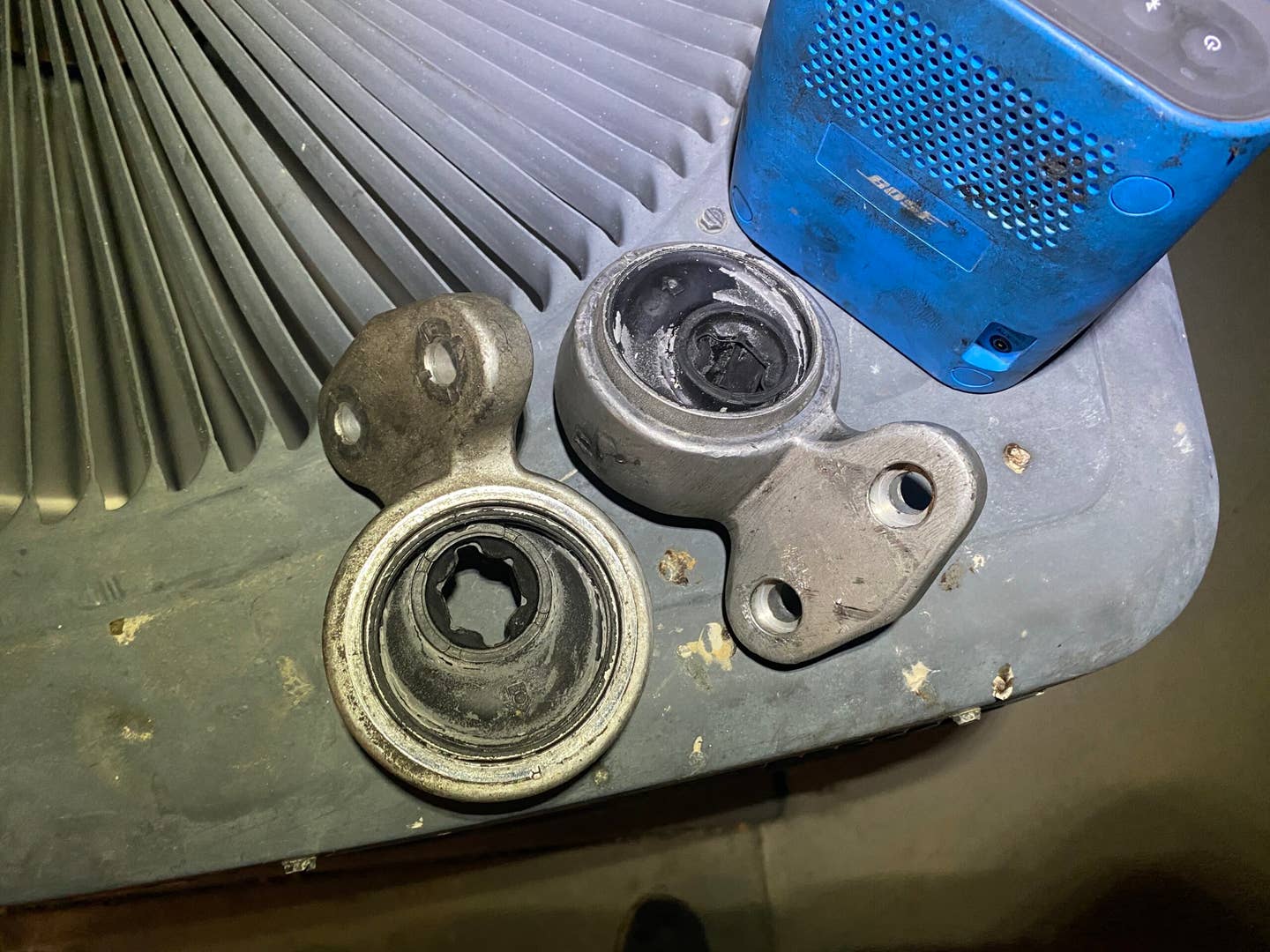 An image of two BMW control arm bushings sitting on a home air conditioning unit. A blue Bluetooth speaker sits just beside of them.