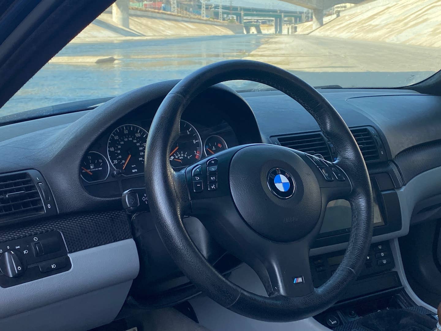 The interior of a BMW 330i ZHP from the open driver's door. The vast concrete expanse of the Los Angeles river stretches beyond the windshield. The lower part of the dashboard is gray.