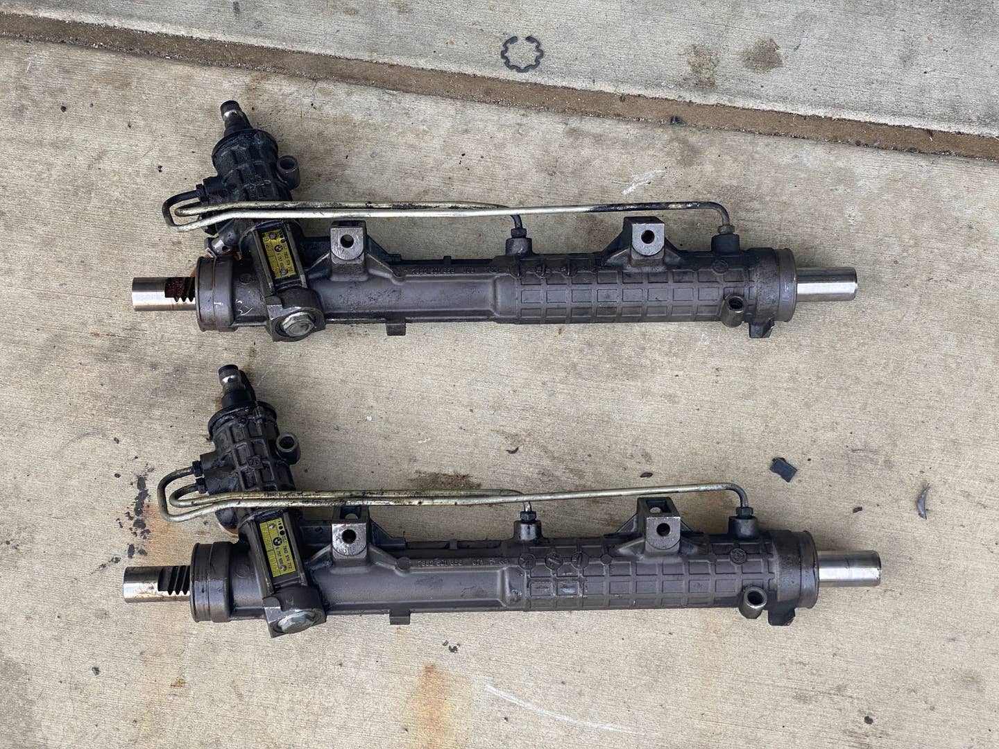 Two steering racks lay side by side on concrete. They both have a gold tag on the input shaft.