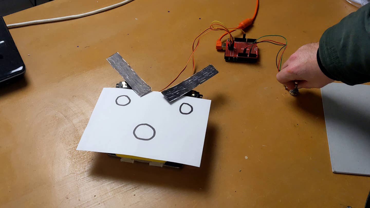 My early proof-of-concept build. The red circuit is the microcontroller board that controls the system. It reads the "throttle position," simulated here with the knob on the right. Based on that, it directs the servos to rotate the eyebrows on this charming cartoon character's face. <em>Lewin Day</em>