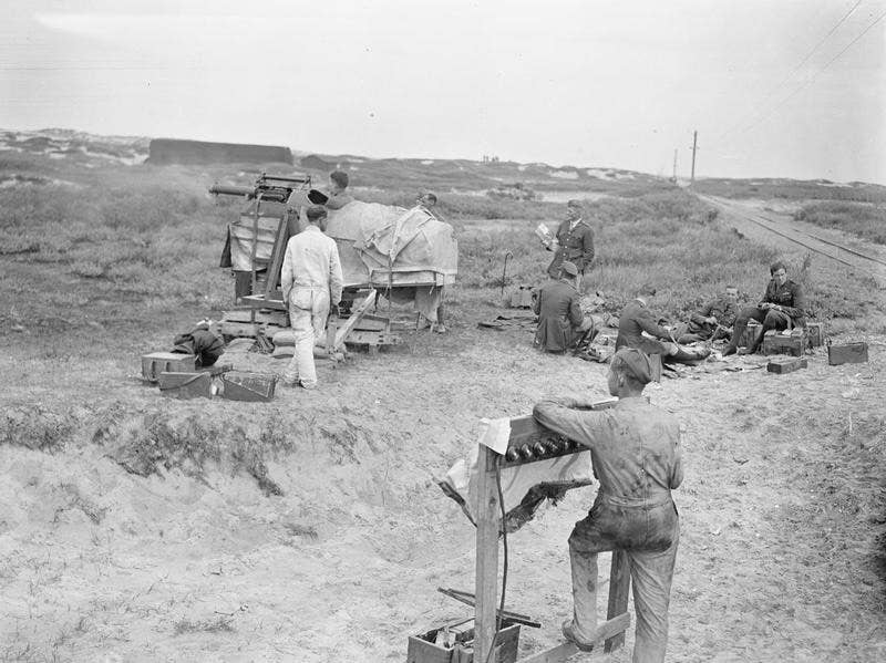 The RAF Gunnery School between Rang-du-Fliers and Verton. A Pilot Officer firing a Vickers gun from a moveable cockpit at targets which disappeared at the end of 6 seconds. The electric controls for this are seen in the foreground. To the right, other Pilot Officers are filling machine gun ammunition belts. July 17, 1918. <em>David McLellan via Wikimedia Commons</em>