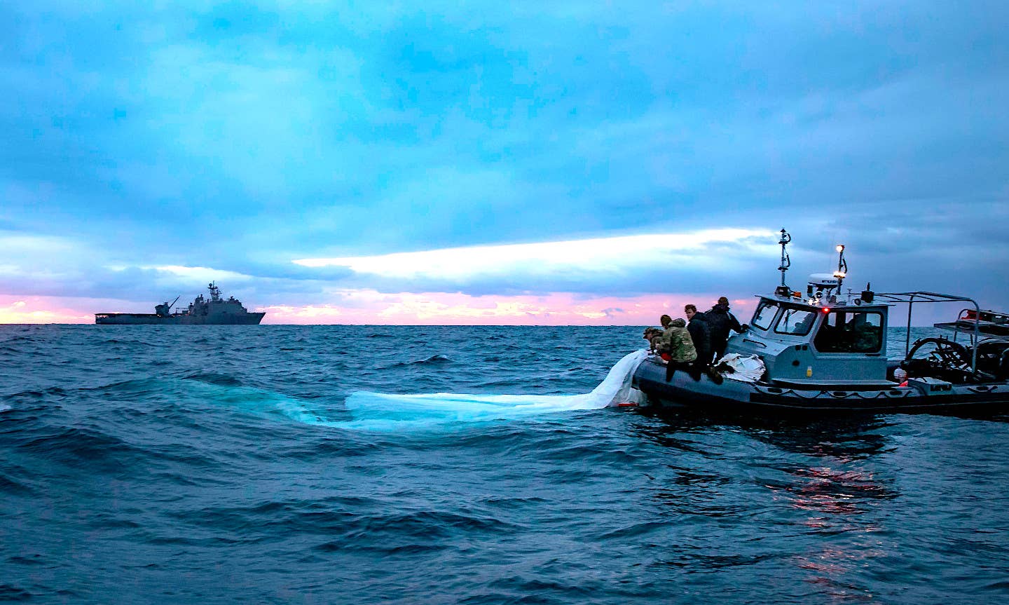 The <em>Harpers Ferry</em> class amphibious warfare ship USS <em>Carter Hall</em> sails in the background as Navy sailors recover a portion of the Chinese spy balloon's envelope. <em>USN</em>