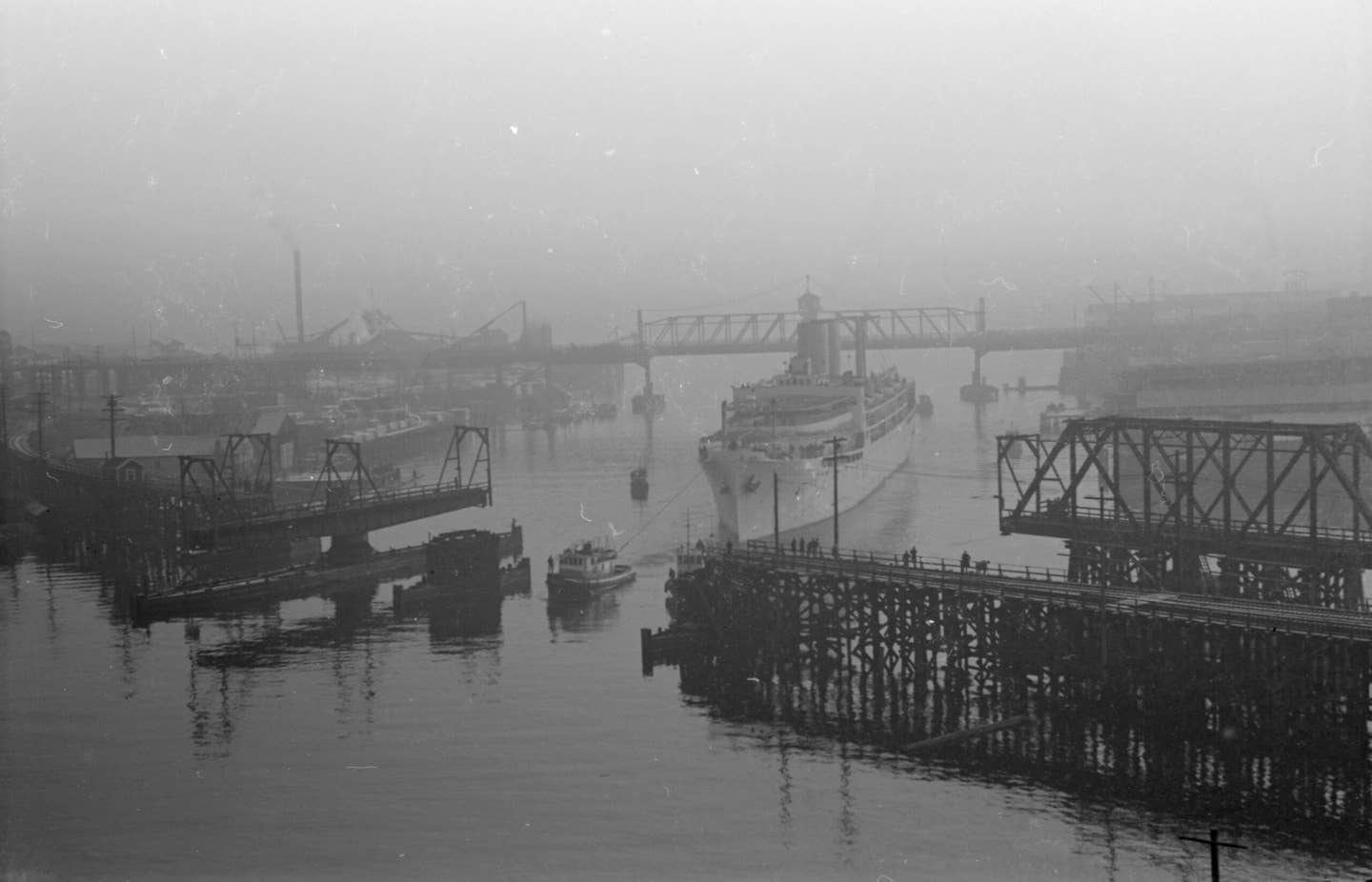 HMS <em>Menestheus</em> being towed out of False Creek toward English Bay by two tugboats on September 1, 1945. Photo likely taken from the south side of Burrard Street Bridge while facing south-east, Vancouver, British Columbia. <em>James Crookall via Wikimedia Commons</em>