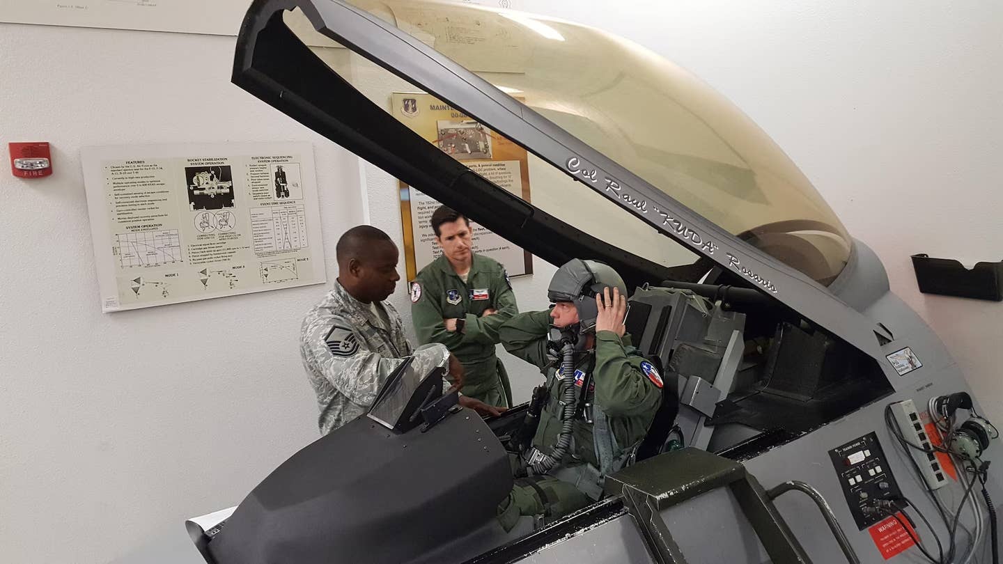 An F-16 pilot sits in a cockpit mockup during egress training on the ground. This training teaches pilots to get in and out of the cockpit safely in various situations, including if they need to eject in an emergency. <em>USAF</em>