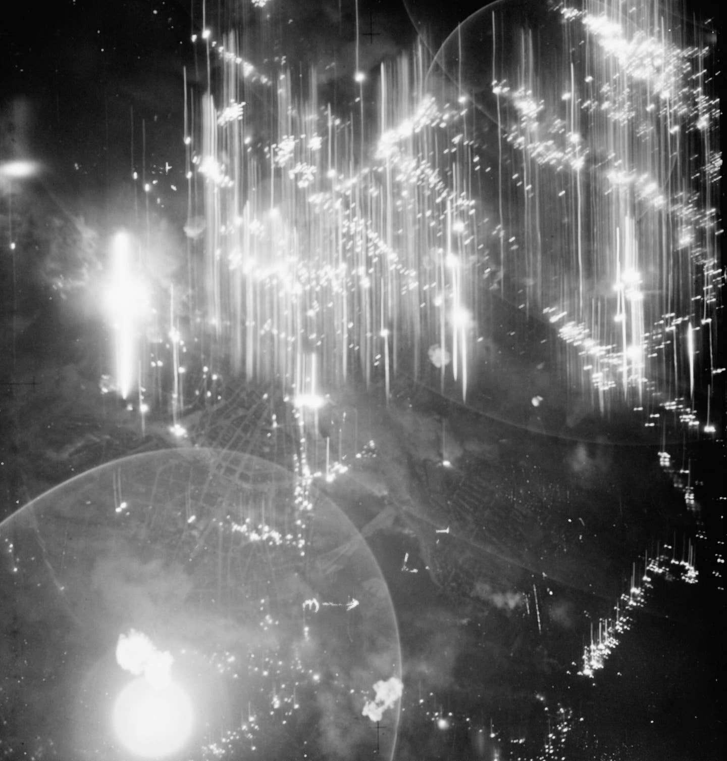 An annotated vertical aerial photo taken during the night raid on Hamburg of July 24/25, 1943. Sticks of incendiaries are seen burning in the Altona and dock districts at the top, while the bright circular object bottom left is a photo-flash bomb. <em>Air Ministry</em>