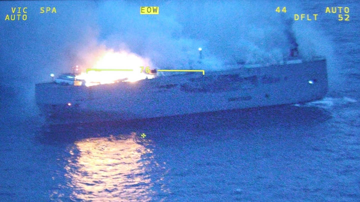 A cargo ship on fire as viewed from the air. A large fire is visibly on the ship's bow, while most of the deck emits smoke.