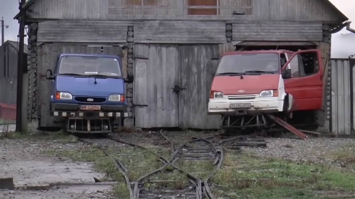 Ford Transit rail vehicles on a narrow-gauge railway in Romania. Both vehicles, one blue and one red with a white bumper, are parked in front of a barn.
