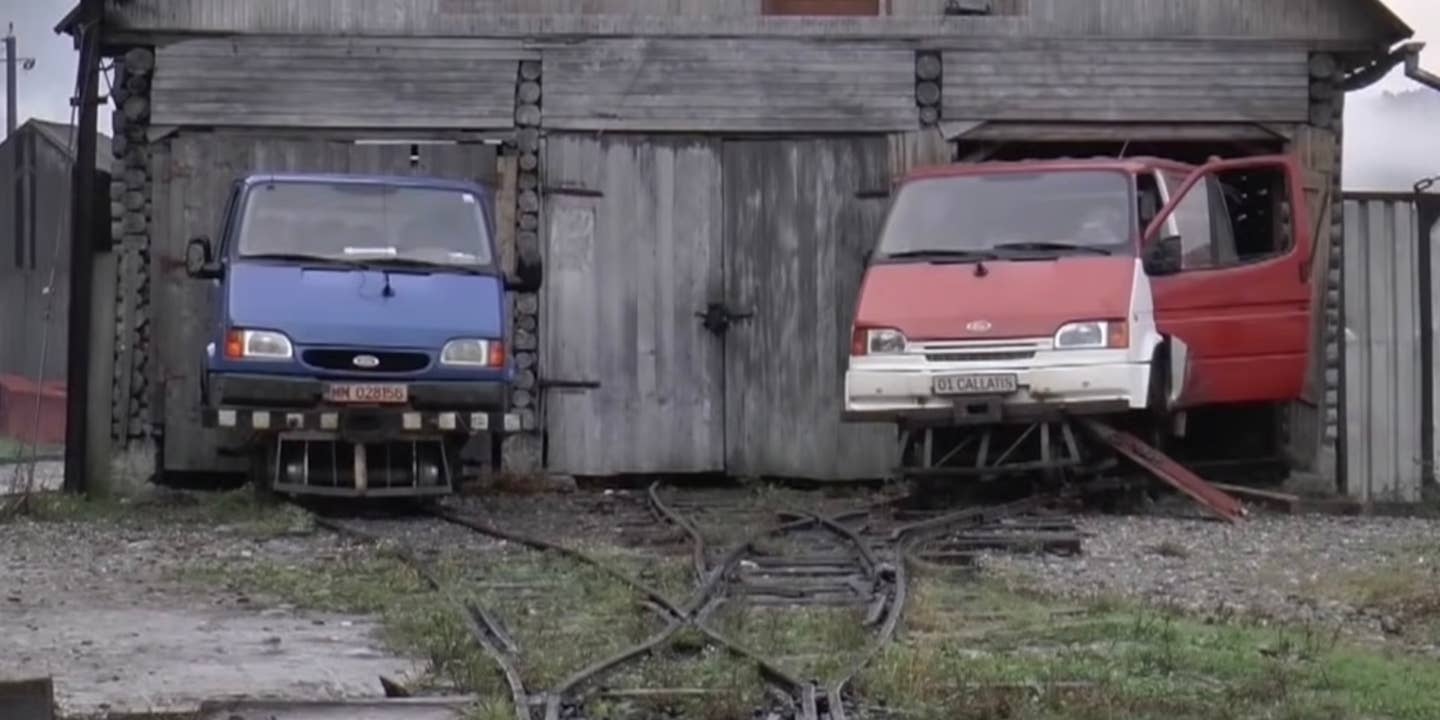 Ford Transit rail vehicles on a narrow-gauge railway in Romania. Both vehicles, one blue and one red with a white bumper, are parked in front of a barn.