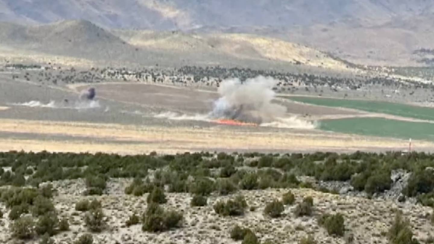 Crash site of fatal Reno Air Races collision. A front of flame can be seen on the desert floor, billowing a large plume of smoke. A second source of smoke can be seen elsewhere.