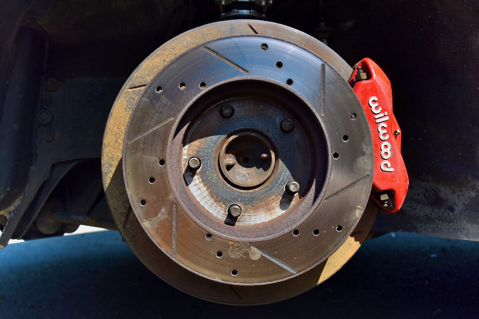 A red Wilwood brake caliper around a large slotted brake rotor, with a smaller drilled and slotted rotor on the front.
