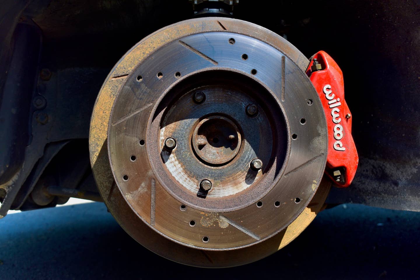 A red Wilwood brake caliper around a large slotted brake rotor, with a smaller drilled and slotted rotor on the front.