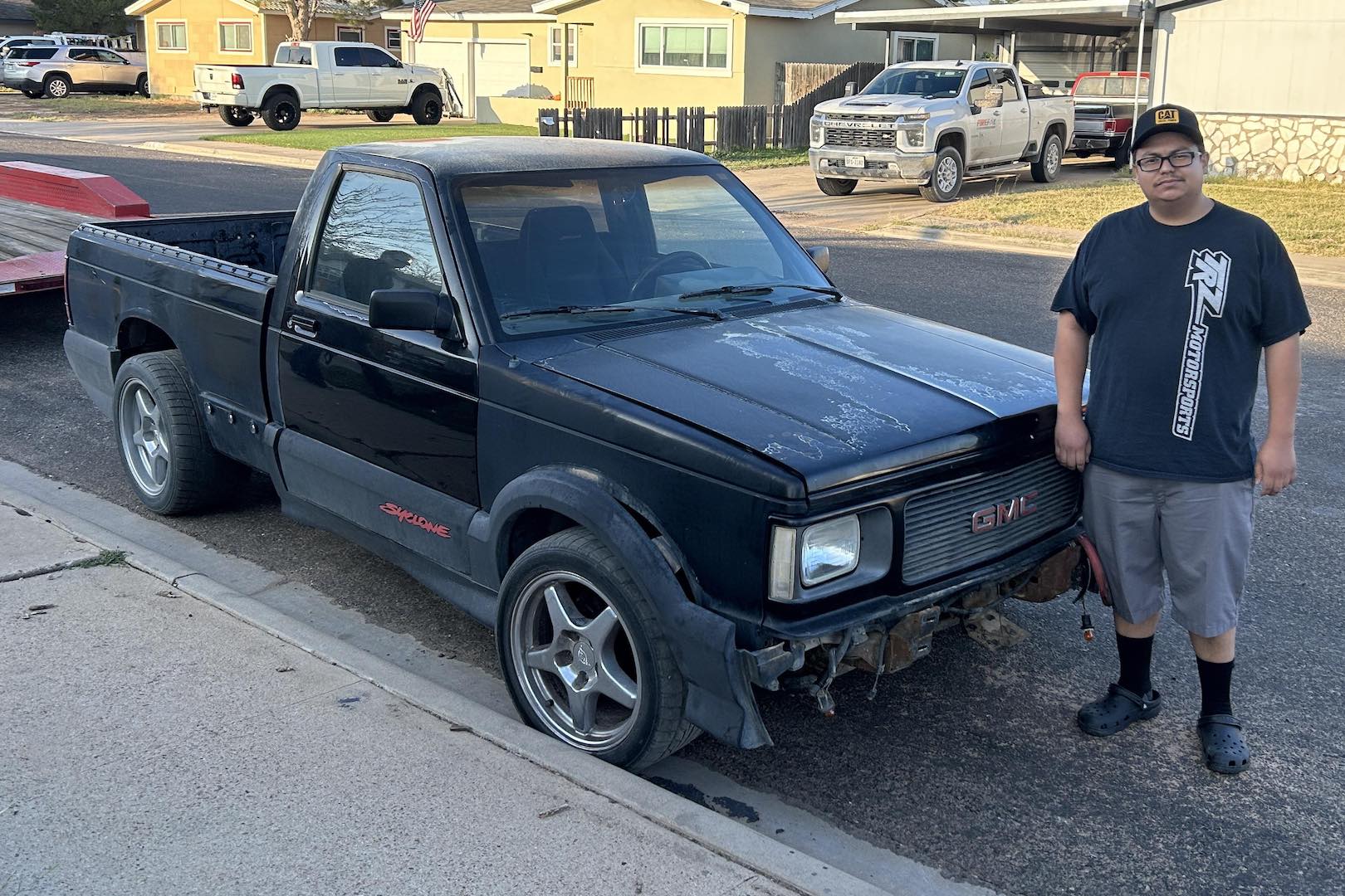 A man in glassed and a cap stands nexts to a black GMC Syclone pickup truck