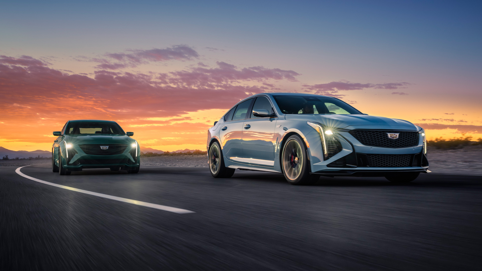 Front passenger view of the 2026 Cadillac CT5-V in Typhoon Metallic (left) and CT5-V Blackwing in Drift Metallic (right) driving at dusk.