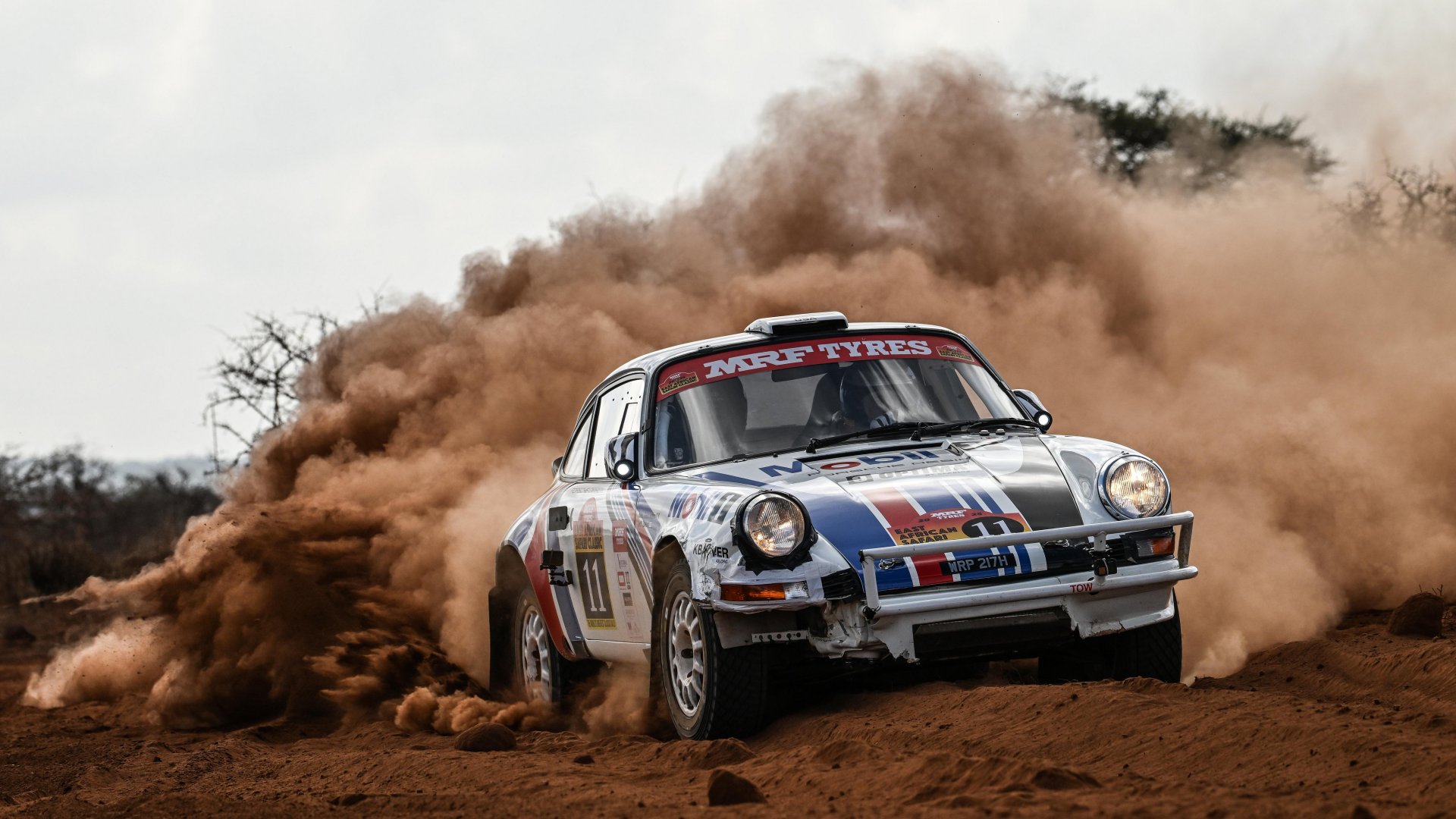 A rally-spec Porsche 911 driving out of a cloud of dust