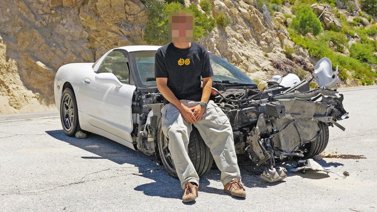 A crashed Corvette and its owner posing with it, with their face obscured.