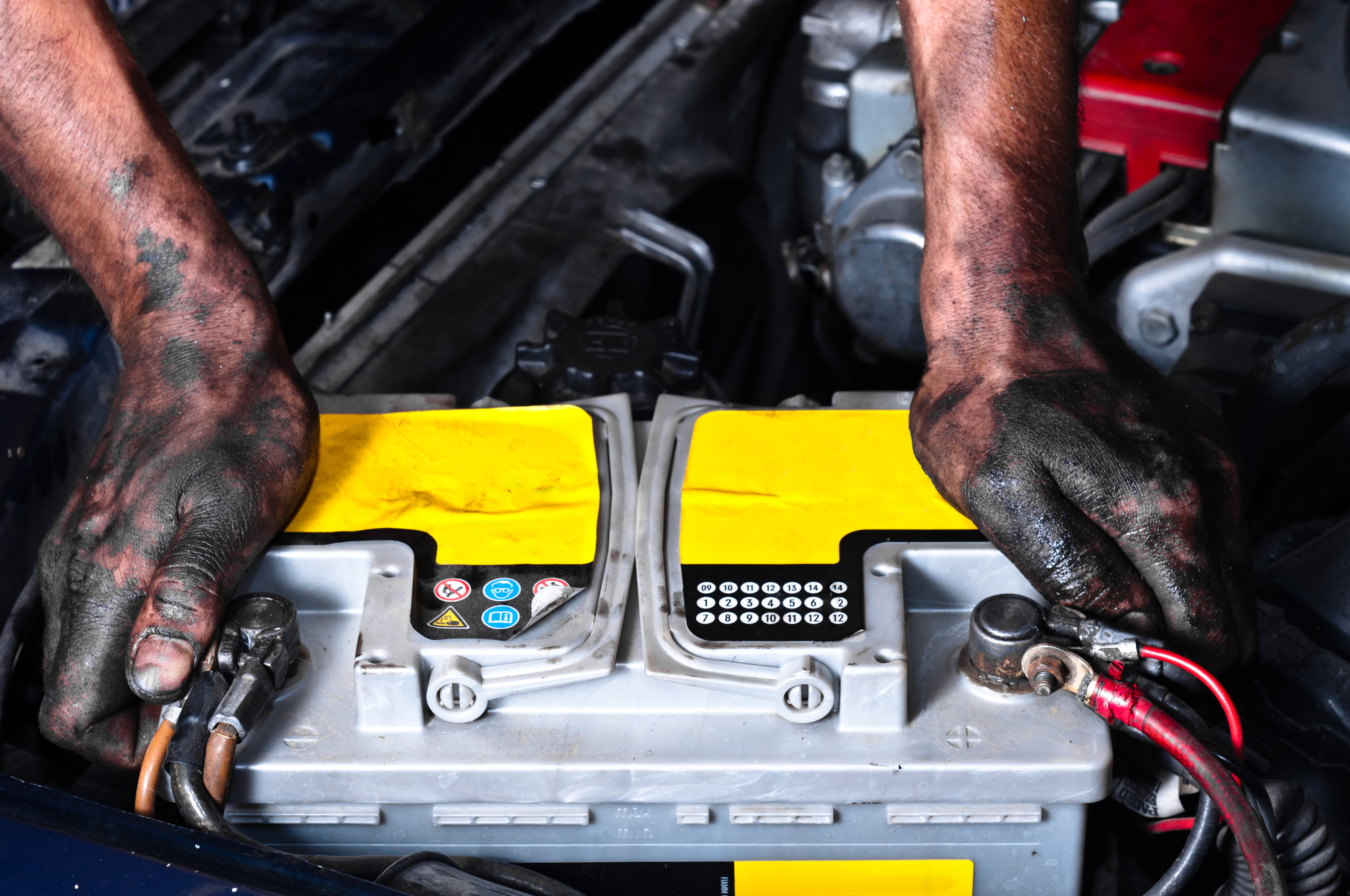 Dirty hands grip a battery in the engine bay.